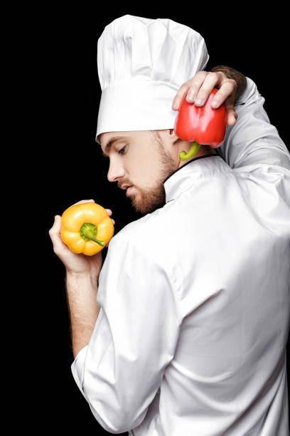 istock Young bearded man chef In white uniform holds bell peppers on  black background 689954620