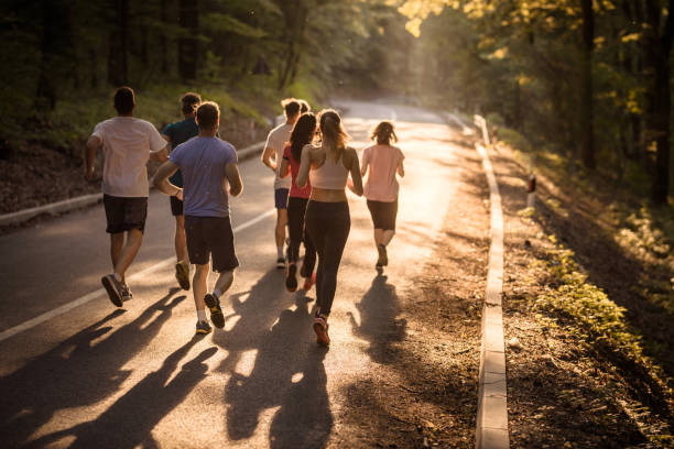 Rear view of marathon runners racing at sunset. Back view of group of people running a marathon on asphalt road at sunset. distance running stock pictures, royalty-free photos & images