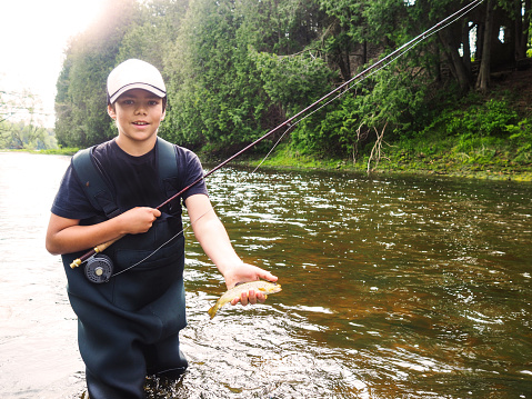 A boy fly-fishing on a river holding a small brown trout he just caught.