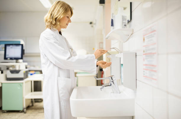 Side view of mature doctor dispensing soap by sink. Female scientist is washing hands at laboratory. She is wearing lab coat.