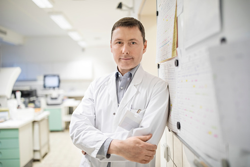 Portrait of confident scientist with arms crossed at laboratory. Mature male doctor is standing by bulletin board. He is in lab coat at hospital.
