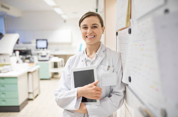 Smiling female scientist holding digital tablet Portrait of smiling young doctor standing by bulletin board. Female scientist is holding digital tablet at laboratory. She is wearing lab coat. technology office equipment laboratory stock pictures, royalty-free photos & images