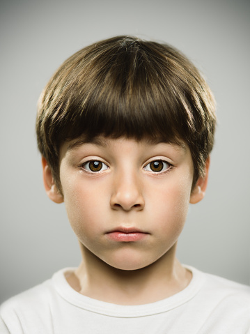 Close up portrait of serious real kid looking at camera. Vertical shot of little boy staring against gray background. Photography from a DSLR camera. Sharp focus on eyes.
