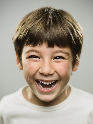 Close up portrait of cute little boy laughing against gray background. Vertical shot of real kid looking excited in studio. Photography from a DSLR camera. Sharp focus on eyes.