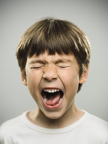 Close up portrait of frustrated little boy screaming against gray background. Vertical shot of real kid shouting with his eyes closed. Photography from a DSLR camera. Sharp focus on eyes.