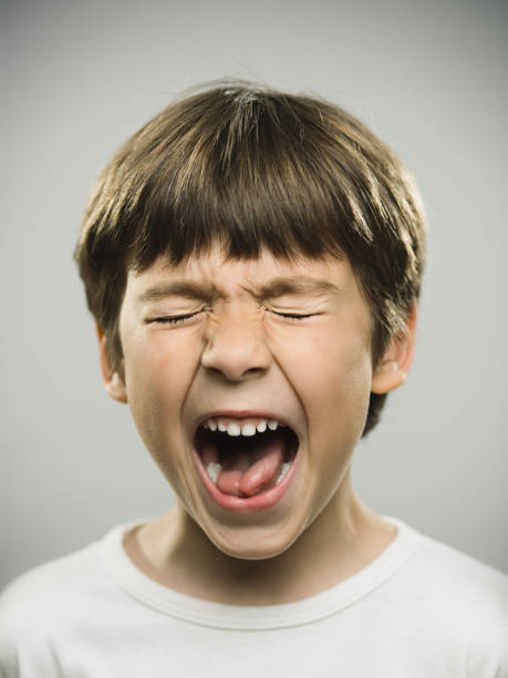 Frustrated little boy screaming Close up portrait of frustrated little boy screaming against gray background. Vertical shot of real kid shouting with his eyes closed. Photography from a DSLR camera. Sharp focus on eyes. irritation photos stock pictures, royalty-free photos & images