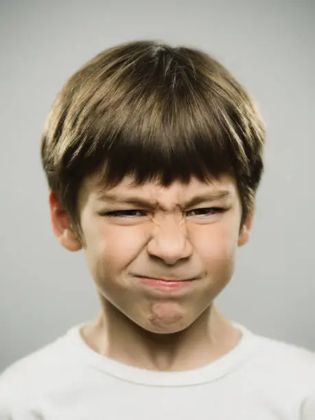Close up portrait of real boy showing disgusted expression in studio. Vertical shot of young kid looking unhappy against gray background. Photography from a DSLR camera. Sharp focus on eyes.
