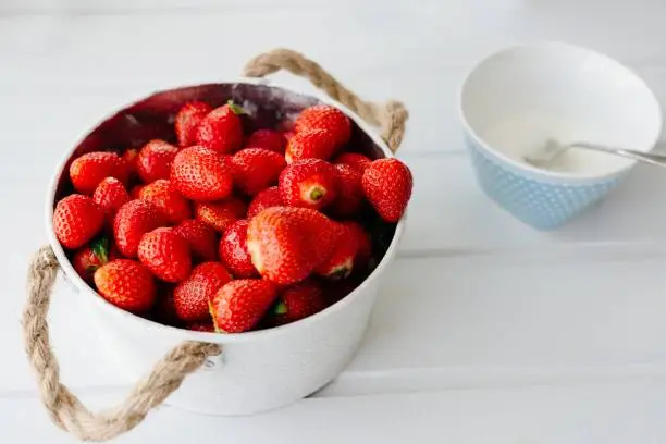 Fresh red strawberries in white bowl and sugarbowl on white wooden table in kitchen
