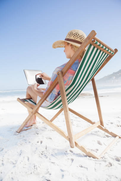 istock Woman in sunhat sitting on beach in deck chair using tablet pc 690035402