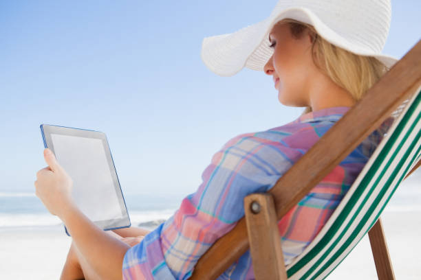 istock Woman in sunhat sitting on beach in deck chair using tablet pc 690035516