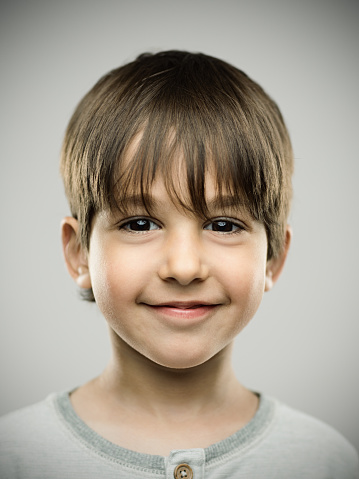 Close up portrait of little boy with sweet smile on his face against gray background. Vertical shot of real kid smiling in studio. Photography from a DSLR camera. Sharp focus on eyes.