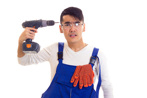 Young serious man with dark hair in white shirt and blue overall with orange gloves and protective glasses holding electric screwdriver on white background in studio.