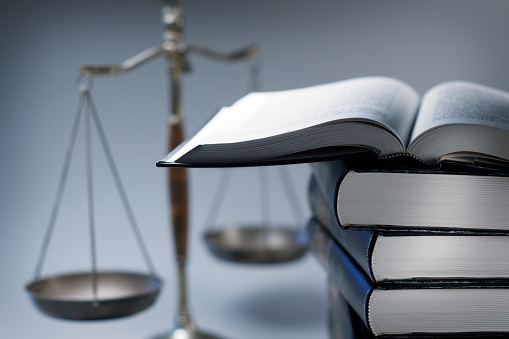 A stack of law books stands in front of a justice scale that is slightly out of focus.  On top of the stack is an open law book.