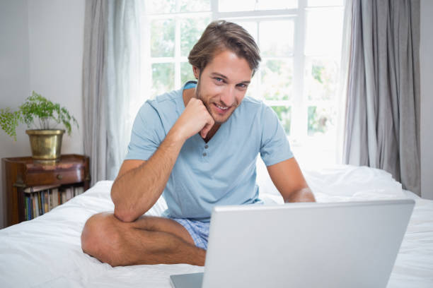 istock Handsome man sitting on bed using laptop smiling at camera 690274620