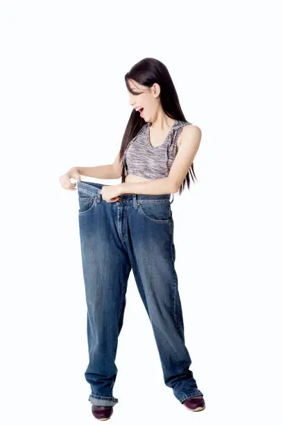 Diet concept. Pretty Asian woman wearing her old jeans and looks happy after success doing diet, isolated on white background