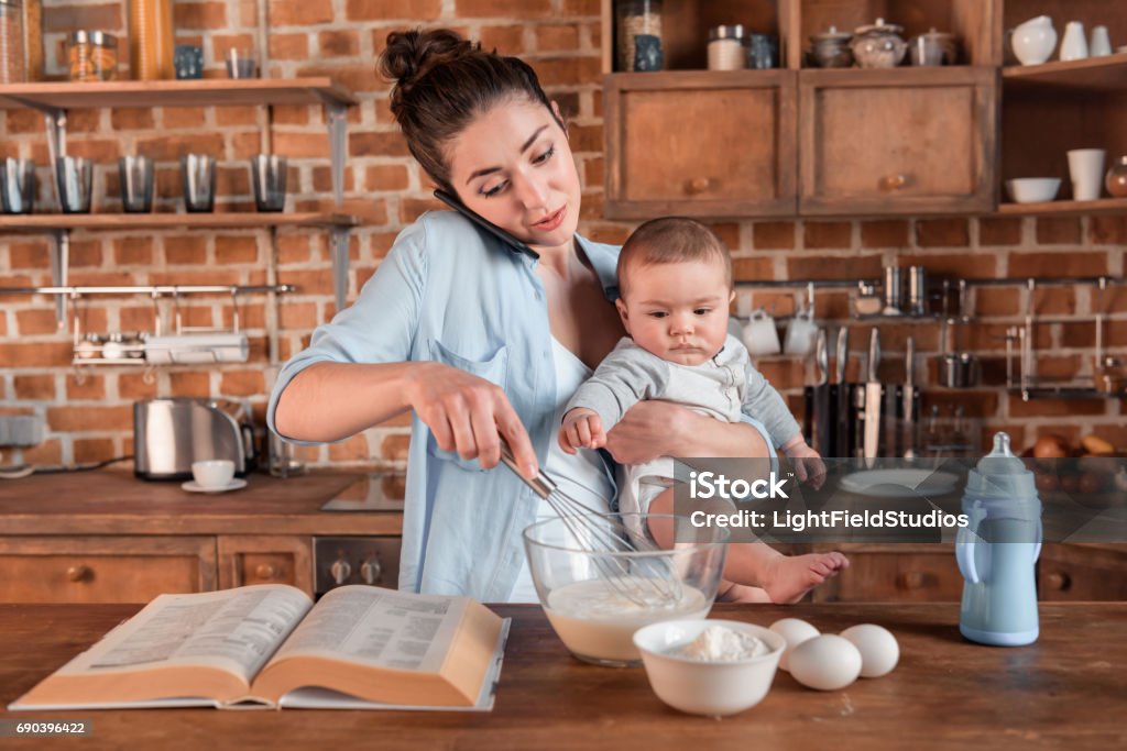 mother holding her son, talking on smartphone and mixing a dough Young mother holding her son, talking on smartphone and mixing a dough at the kitchen. family life and multitasking concept Mother Stock Photo