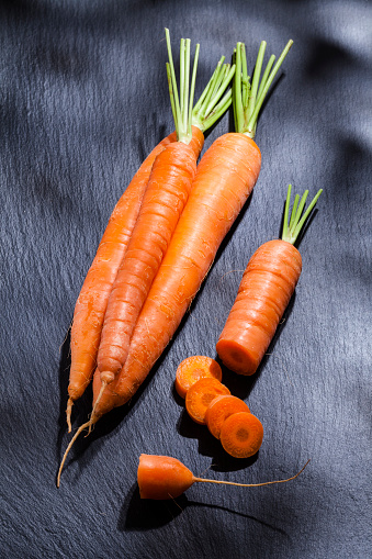 High angle view of four organic carrots on black slate table. One carrot is cut into slices. Predominant colors are orange and black. Low key DSRL studio photo taken with Canon EOS 5D Mk II and Canon EF 100mm f/2.8L Macro IS USM