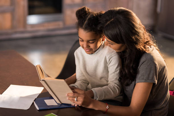 istock Smiling mother and daughter sitting together at table and reading book, homework help concept 690412140