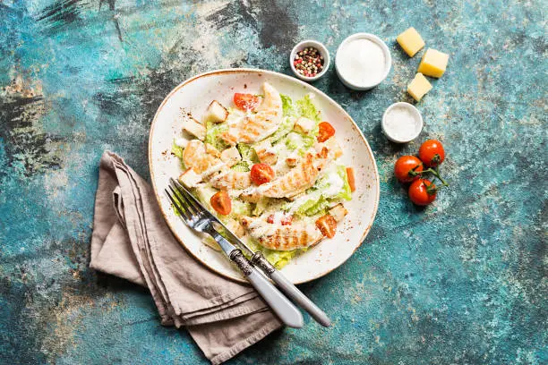 Delicious caesar salad with croutons, grilled chicken breast, grated parmesan cheese and cos lettuce, with sauce in the gravy boat, simply and healthy recipe, top view
