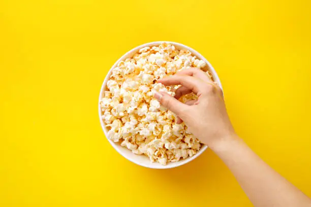 Photo of Popcorn viewed from above on yellow background. Woman eating popcorn. Human hand. Top view