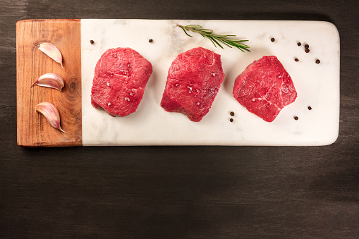 Three slices of raw meat, beef fillet, shot from above on with a sprig of rosemary, garlic cloves, salt, and pepper, with a place for text