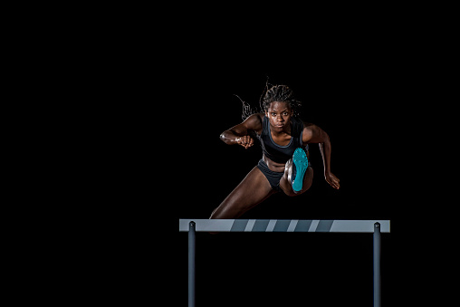 Front view of a female african-american athlete jumping over a hurdle. Black background.