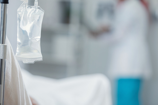 Close up of iv drip bag in a patient's hospital room. A healthcare professional is blurred in the background.