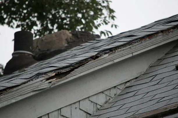 Roof of a residential house showing damage to the tile and gutter systems, multiple layers of shingles, missing shingles, and rusty gutters.