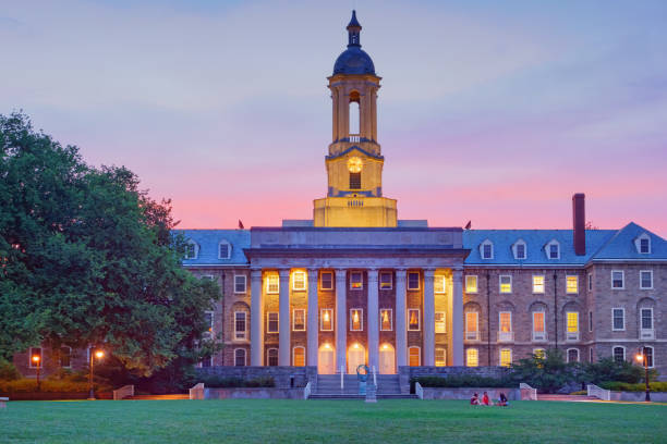 Penn State Old Main Building in State College Pennsylvania USA Students chat in front of Old Main, the main administrative building of Penn State after sunset. It is located at University Park in State College, Pennsylvania, USA. Pennsylvania State University (commonly referred to as Penn State or PSU) is a public university with campuses and facilities throughout Pennsylvania. old main penn state stock pictures, royalty-free photos & images