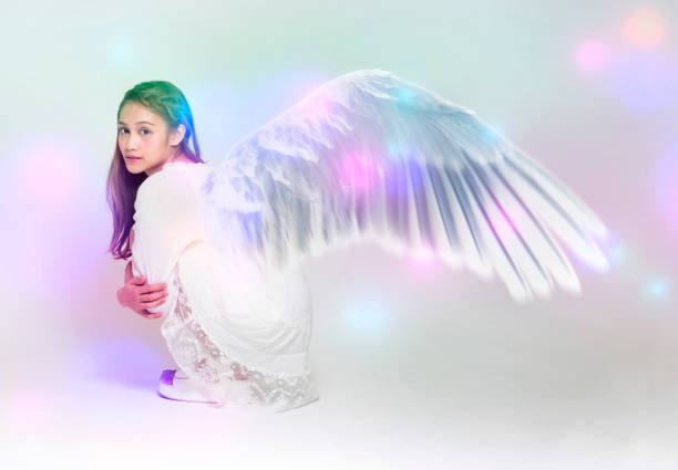 Cute blonde in fairy costume with wings and lace, transparent white dress sits in front of waterfall. Fairy wings are golden, made of transparent chiffon, decorated with embroidery. Concept of magic