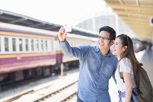 Young couple asian woman and man traveler are looking the smartphone for selfie themself with train station background. Travel in summer concept.