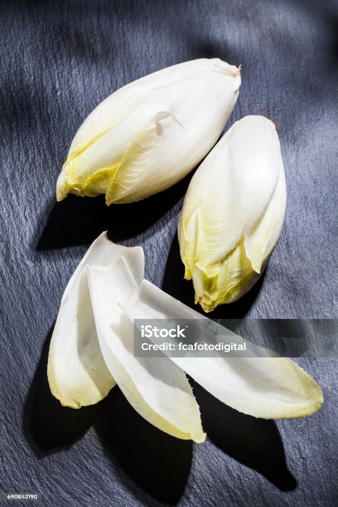 Endives on black slate background High angle view of two whole organic endives on black slate table. Three endive leaves are on foreground. Predominant colors are white and black. Low key DSRL studio photo taken with Canon EOS 5D Mk II and Canon EF 100mm f/2.8L Macro IS USM Endive Stock Photo