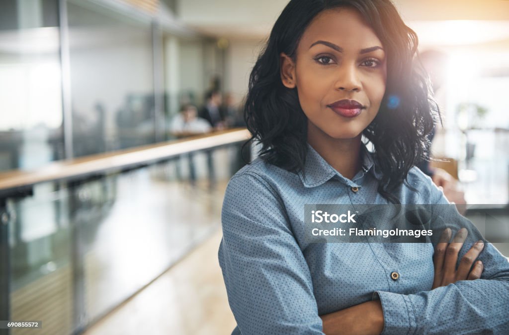 Beautiful black businesswoman with arms crossed in office Pretty young dreamy African-American office worker standing with arms crossed and looking at camera. Businesswoman Stock Photo