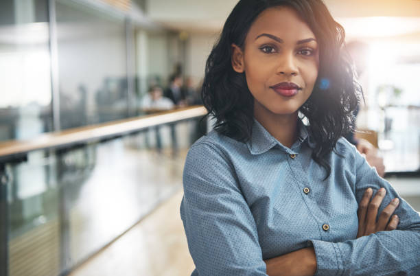 Beautiful black businesswoman with arms crossed in office Pretty young dreamy African-American office worker standing with arms crossed and looking at camera. confidence stock pictures, royalty-free photos & images
