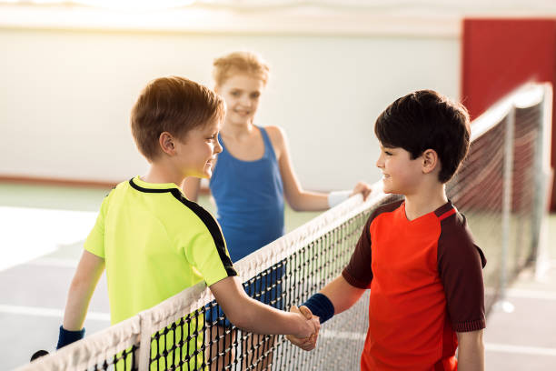 Good game. Joyful two boys and shaking hands after playing tennis. They are looking at each other and smiling. Girl is standing on background
