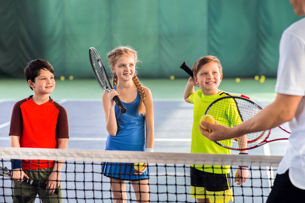 Joyful pupils learning to play tennis Excited children are listening to explanations of their tennis trainer with attention. They are holding rackets and laughing. Portrait tennis coach stock pictures, royalty-free photos & images