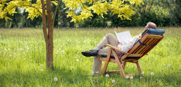 istock Relaxing Senior man lying in chair on meadow under tree reading book 690893970