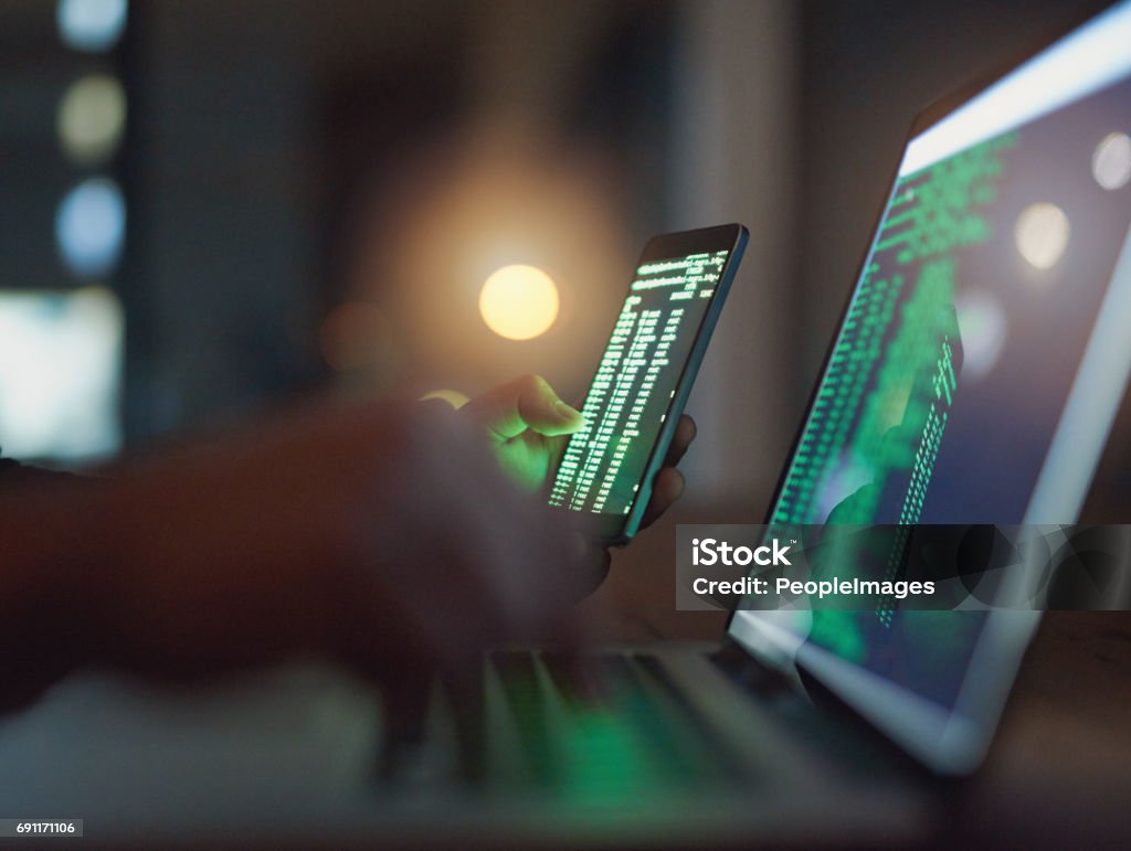 Who knows what a hacker can do with your information Shot of an unrecognisable hacker using a cellphone and laptop in the dark Network Security Stock Photo