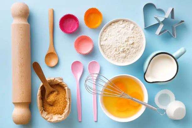 Photo of Baking utensils and ingredients. Egg yolk, brown sugar, milk, flour, whisker, spoons, cinnamon, bowl, rolling pin, cupcake paper cup, molds, sweet decoration elements. Top view. Flat lay