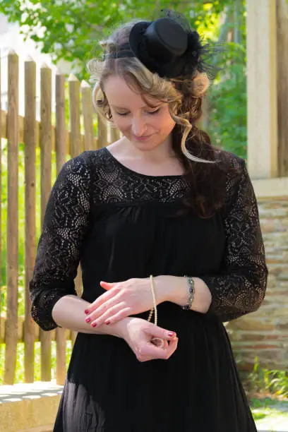 Woman in vintage black dress and small hat trying on a bracelet standing outside near wooden fencing and looking down at her wrist