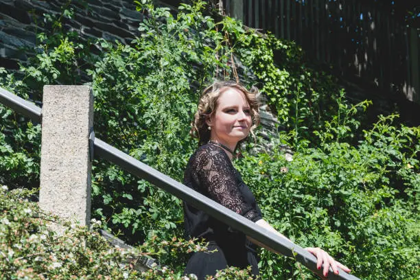 Pretty young woman in a stylish lacy black dress standing on an outdoor staircase amongst overgrown greenery looking ahead with a smile and dreamy expression