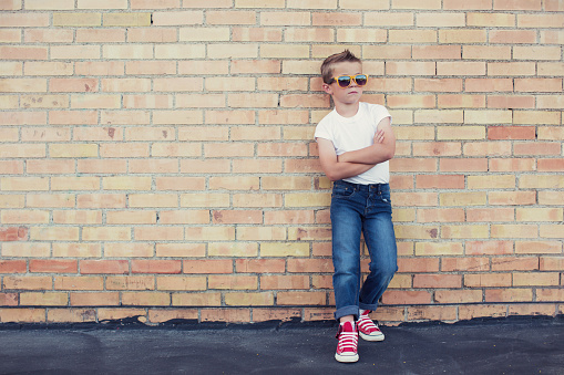 A young boy dressed as a rebel in white t-shirt, sunglasses and jeans with faux hawk folds his arms while looking cool and rebellious. Image taken in an alleyway in front of a brick wall.