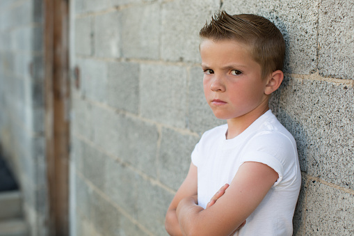 A young boy dressed as a rebel in white t-shirt and jeans with faux hawk folds his arms while looking sternly and rebellious at the camera. Image taken in an alleyway in front of a brick wall.
