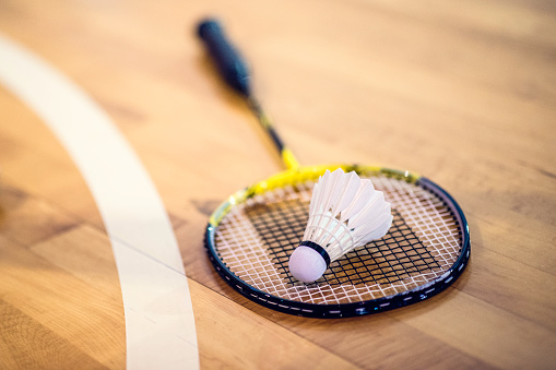 Close-up shot of a badminton racket and a shuttlecock on a wooden court flooring.
