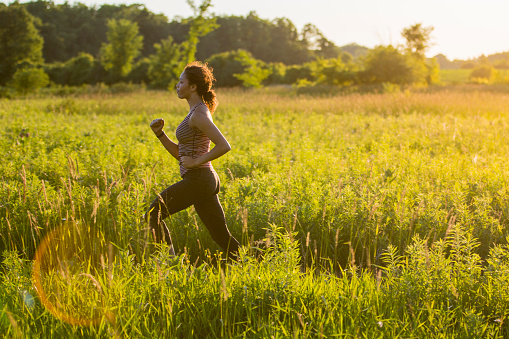 A woman of African descent is outdoors. She is in a grass field during springtime. She is wearing athletic clothing. She is jogging through the field during sundown.