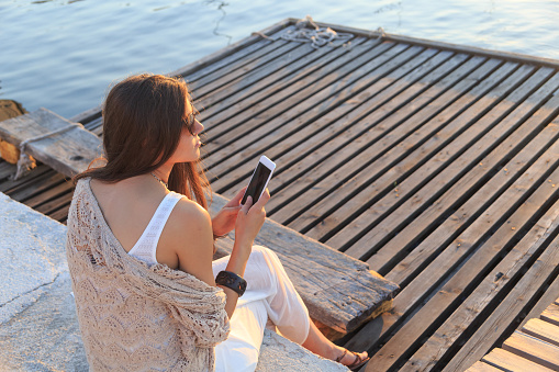 Young woman sitting on beach and using smart phone. Wears casual off shoulder top, with sunglasses, long hair.