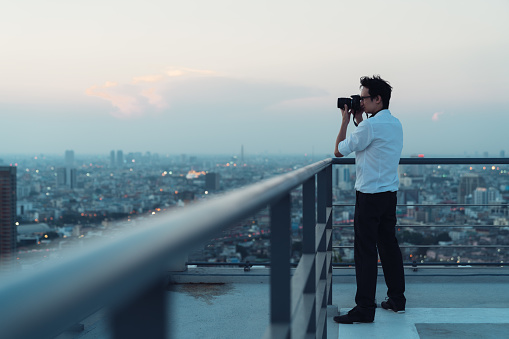 Asian man taking cityscape photo on building rooftop in low light situation. Photography, office people, or hobby concept. With copy space