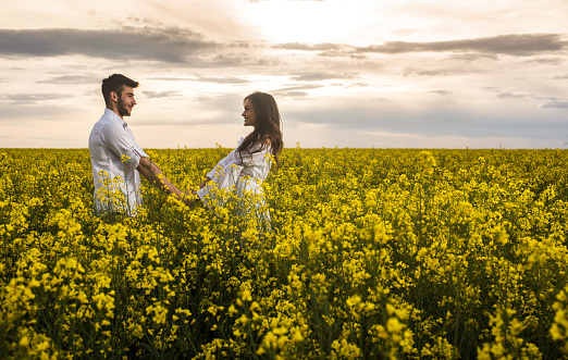 Happy couple in love holding hands in a field of yellow flowers and looking at each other.