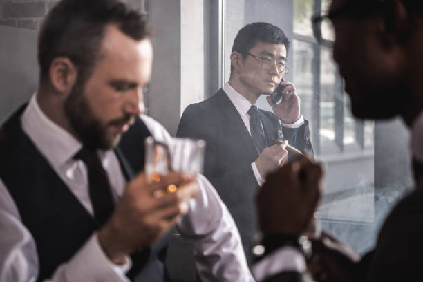 istock Serious asian businessman smoking cigar and talking on smartphone while colleagues drinking whisky, multicultural business team 691429436