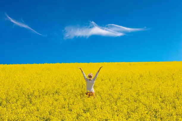 Outdoor shot of a happy girl in rapeseed field with arms raised up to blue sky